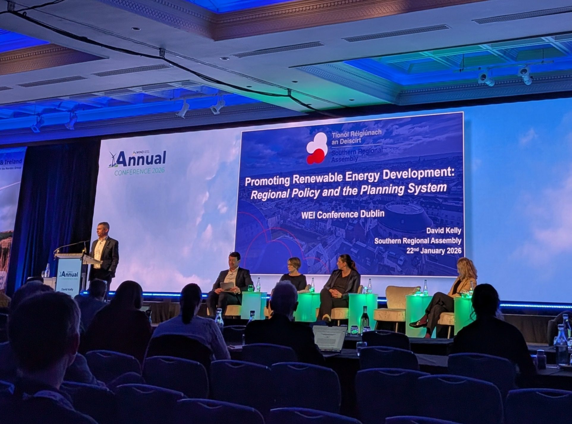 View of four panelists sitting on stage and speaker David Kelly, Director of the Southern Regional Assembly presenting at a podium. 