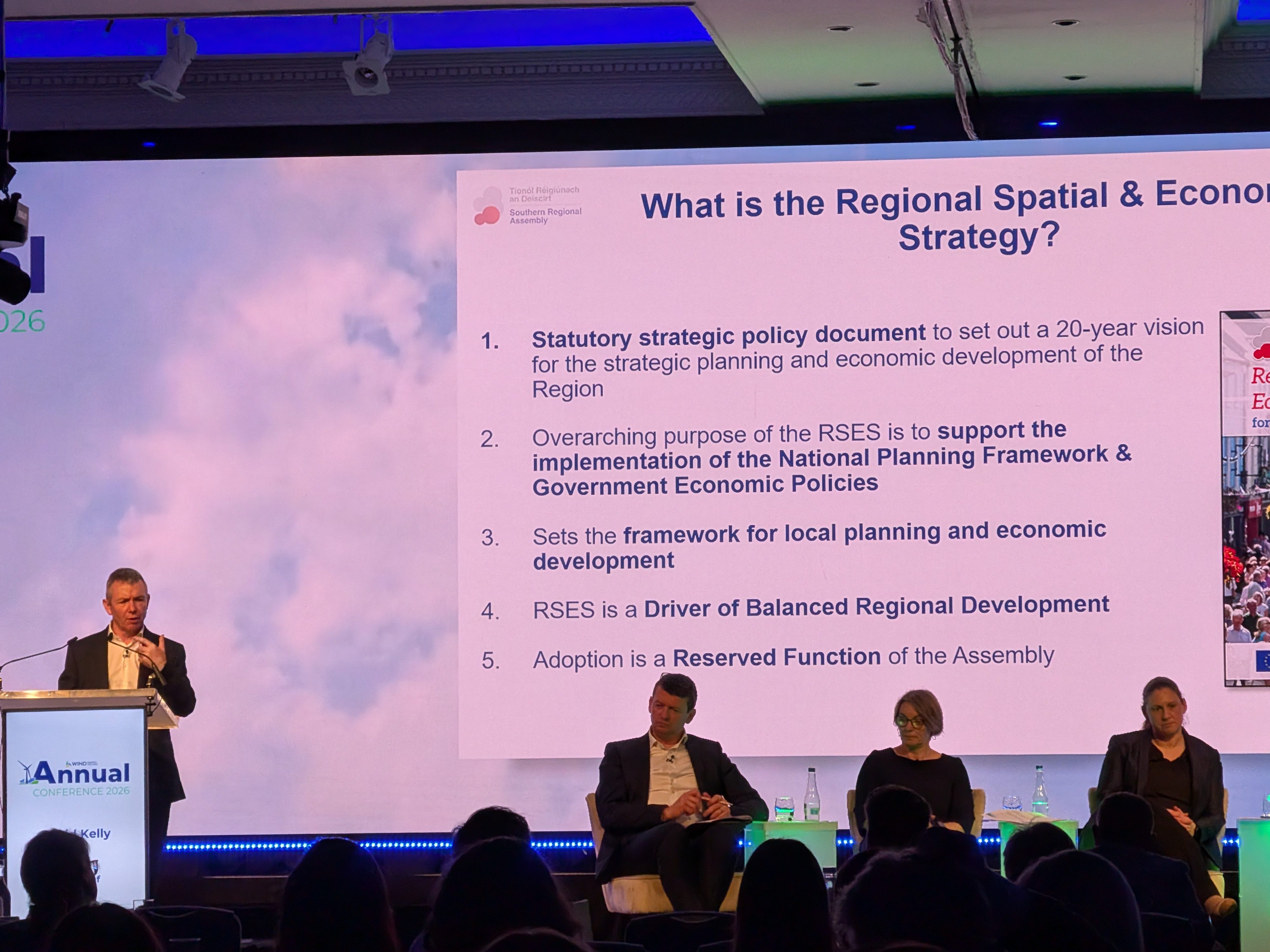 David Kelly, Director of the Southern Regional Assembly presenting at a podium to an audience. Three others are also on stage sitting down.